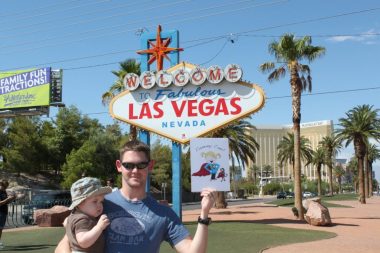 Cammy Can | Rett Syndrome News | A young father in sunglasses and a T-shirt holds a toddler in front of a "Welcome to Las Vegas" sign, and he holds a printout of the Cammy Can cartoon figure 