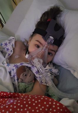 911 | Rett Syndrome News | Cammy lies in a hospital bed hooked up to a BiPAP machine and holding a Curious George stuffed animal