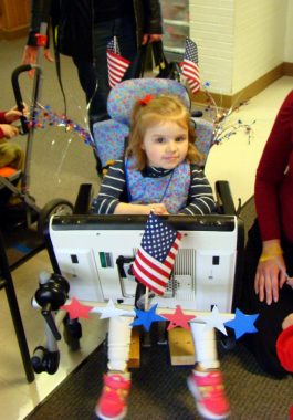 communicating with Rett | Rett Syndrome News | Cammy sits in her wheelchair with her eye-gaze communication device propped in front of her. She is wearing red, white, and blue clothing, and her equipment is decorated with American flags and stars in honor of Veterans Day.