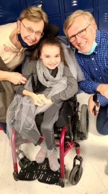 school play | Rett Syndrome News | After the school play, Cammy smiles between her grandmother and grandfather. She is wearing gray and white tulle for her role as a tornado, and is sitting in a pink wheelchair.