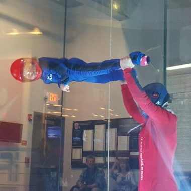indoor skydiving | Rett Syndrome News | Cammy hovers inside an indoor skydiving chamber parallel to the ground as an instructor holds on to her feet. Cammy is wearing a blue, full-body suit and a red helmet.