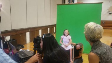 picture day | Rett Syndrome News | In front of a green screen, a smiling Cammy sits on a box. Behind her a woman on her knees has a hand supporting her back. Another woman, in the right foreground, is drawing Cammy's attention. In the center foreground is a photographer, with a camera in front of her and a laptop nearby.