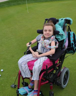 golf | Rett Syndrome News | Cammy in her wheelchair on a putting green, with a club at her side