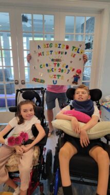 Two 14-year-olds, one girl and one boy, sit side by side in their respective wheelchairs after a homecoming proposal. The boy holds a pillow in his lap while the girl holds a small bouquet of pink flowers. The boy's father stands behind them holding up a sign that reads, "A big bird told me it would be super cool to go to Hoco 2023 with u" in multicolored letters. The sign is decorated with Big Bird from "Sesame Street" and the Super Mario Bros.