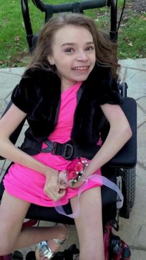 A girl sits in her wheelchair on a gray outdoor patio; green grass is in the background. She has long brown hair and wears a short and bright pink dress, a red wrist corsage, and a black sweater or jacket.