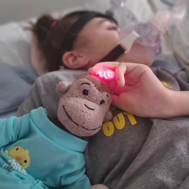 A child lies in bed with a plastic mask over her face and a stuffed monkey at her side. The child is in a gray T-shirt, and her hair is tied up in a bun.