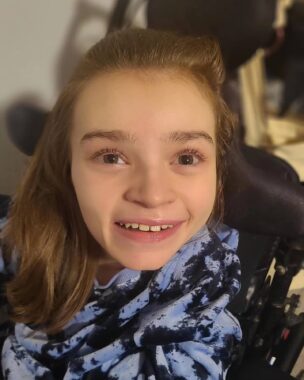 A young girl with Rett syndrome looks up and flashes a bright smile to the camera. Her eyes are wide open and full of light. 