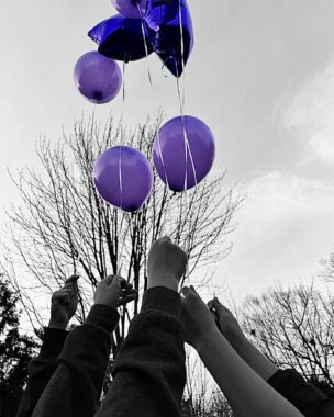 Five arms reach toward the sky holding balloons. The balloons are purple, and so they stand out in the otherwise black-and-white photo.