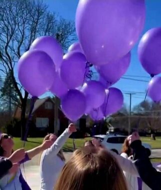 A group of people gather outside in front of the author's house on a sunny day. They are holding purple balloons up toward the sky.