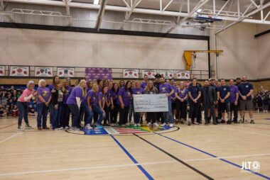 A photo of a big group of people in a school gymnasium. The photo is taken from across the gym, so you can't see anyone's face clearly, but most of them are wearing purple T-shirts, and some of them are holding up a giant symbolic donation check.