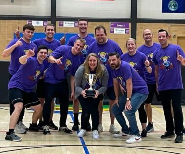 Eleven people pose on the floor of a school gym. They are all wearing purple T-shirts. Some make the No. 1 sign with their hands, while a woman in the center of the photo holds a trophy. 