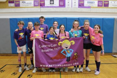 A blurry photo taken in a middle school gym shows 10 seventh graders holding up a large purple banner with a cartoon girl in pony tails and text that reads "Cammy Can Reverse Rett." 