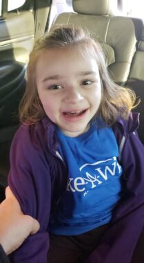 A young girl, about 9 years old, gives a big smile while seated inside a limo. She's wearing a blue Make-A-Wish T-shirt and a purple jacket, and it looks like someone is holding on to her arm.