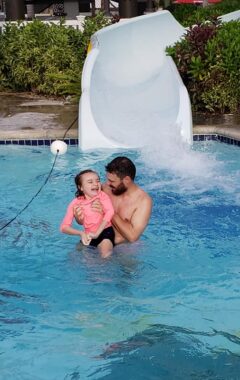 A girl laughs as her dad holds her up in a pool next to a water slide. She's wearing a pink shirt and black shorts, and she and her dad are smiling at each other.