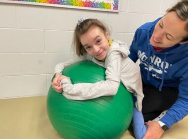 A young girl with Rett syndrome has physical therapy on a large, green ball. She looks up and smiles at the camera. Behind her, holding her legs and looking at her smiling, is a woman in a blue hoodie, who is the girl's physical therapist. 