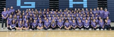 In a gymnasium, at least five lengthy rows of people in identical purple T-shirts sit, kneel, or stand.