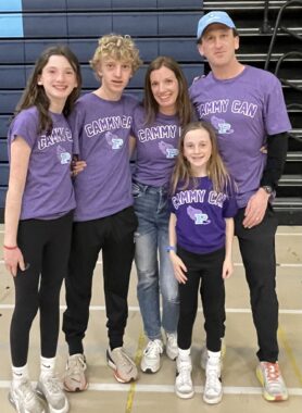 What appears to be a family of five stand in a gymnasium, all in identical purple T-shirts. The boy and girl on the left appear to be teens, with an adult woman and man standing beside them. A preteen girl in in the front between the two adults.