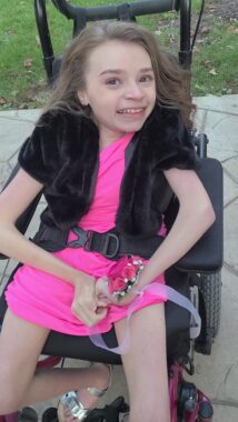 A teenage girl smiles for a photo on homecoming night. She's wearing a pink dress with a black shrug and is holding a corsage with pink flowers. She's seated in her power wheelchair, which looks to be parked on a patio outside her home.