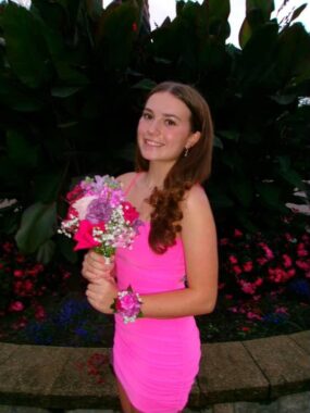 A teenage girl poses for a photo on homecoming night wearing a pink dress and a corsage and holding a bouquet of flowers. The background is dark, but she looks to be standing outside in front of a bed of flowers.