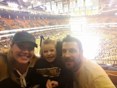 A woman, young daughter, and husband take a selfie in the upper deck of a large hockey arena. They're all smiles. 