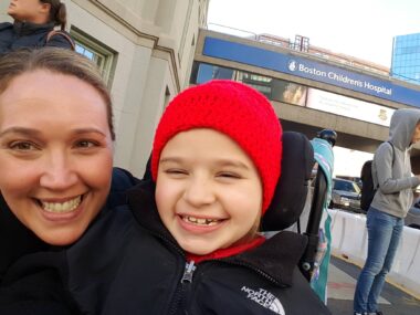 A woman and her young daughter smile broadly in a selfie outside a hospital. They're bundled up for chilly weather. 