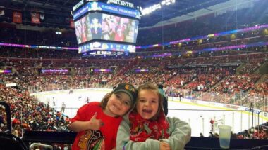 Two young children decked out in the red clothing of the Chicago Blackhawks smile elatedly, while one gestures with a thumbs up, in front of a large ice rink and packed stadium. 