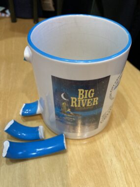 A white, ceramic mug with the "Big River" logo sits atop a wooden table. Its blue handle lies next to it in several pieces.