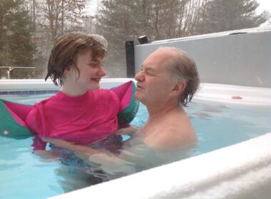A man and his adult daughter enjoy a heated swim spa in their backyard while the snow falls around them. 