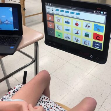 An eye-gaze system is show, with the legs of a girl to the bottom left of the frame, indicating that the girl is looking at the screen. The screen has various folders on it, similar to the home screen of any Microsoft software program.