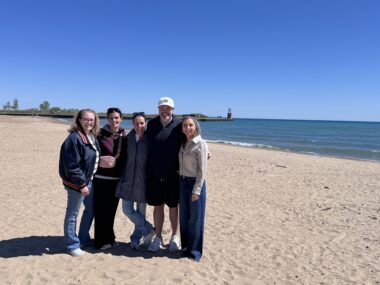 Four women and one man stand on a beach, with jackets to ward off the early springtime cold, on a sunny day, with a cool, calm, and blue lake in the background. The sky is a bright, cloudless blue. 