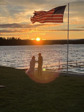 Two people are silhouetted beneath an American flag as they stand near a pier that juts out onto a lake at sunset.
