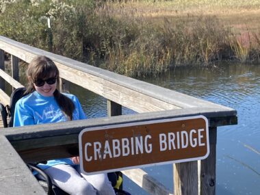 A young woman in dark sunglasses and a blue sweatshirt sits on a woodn bridge or dock over a wetland, behind a sign that says "crabbing bridge."