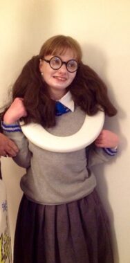 A young woman is dressed up in older clothing from the Harry Potter Films, with round black glasses, her hair in two pony tails, and a toilet seat around her neck.