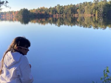 A view from behind a young woman in a white hoodie who is looking to her right. In front of her is a large body of blue water reflecting the sky, and a line of trees also reflected in the water. 